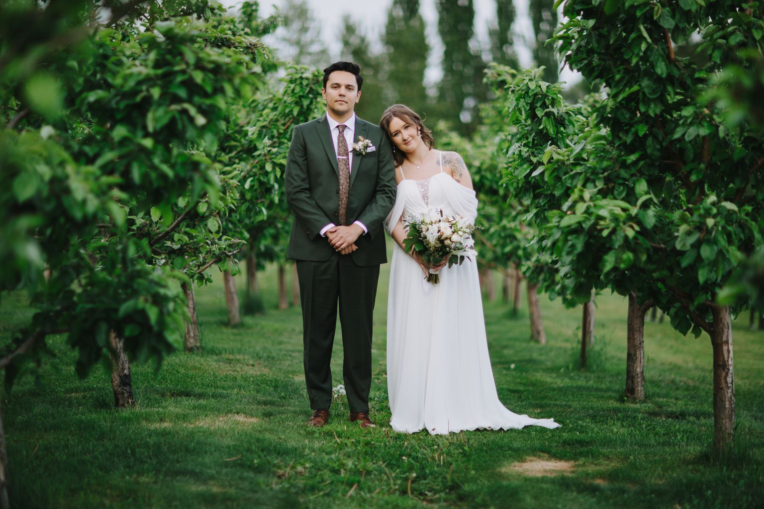 Bride in flowing white gown and groom in dark suit stand together on grass with evergreen trees and soft natural light behind them