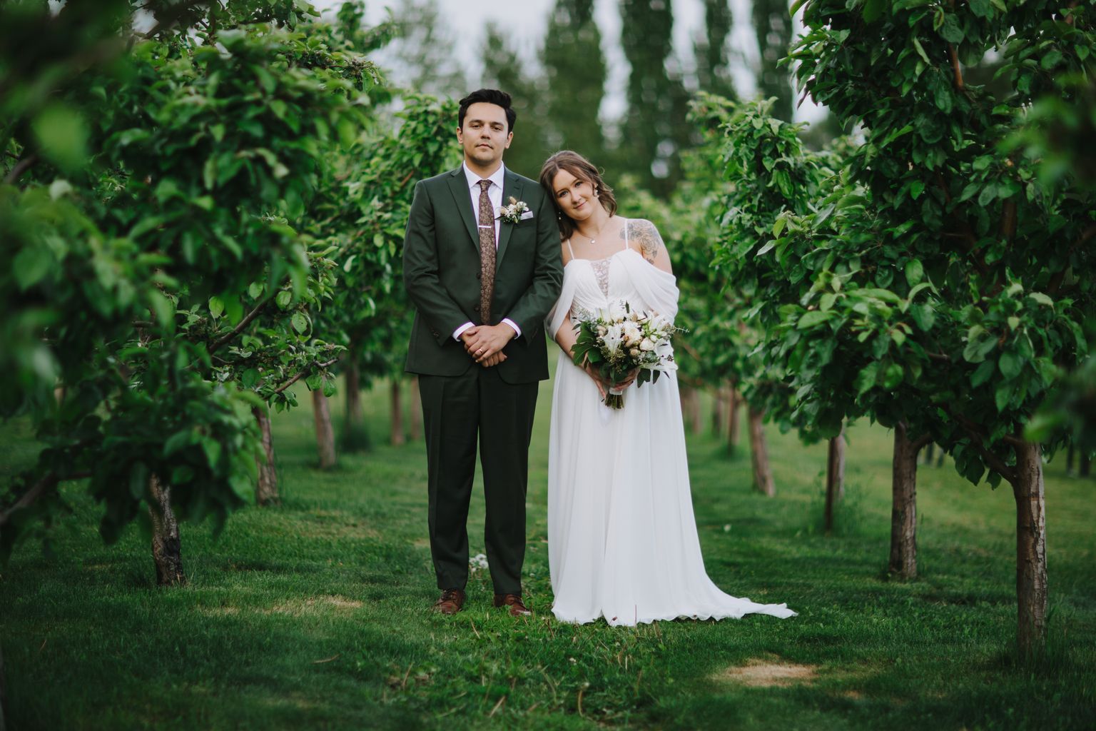 Bride in flowing white gown and groom in dark suit hold hands in lush garden setting with tall evergreen trees