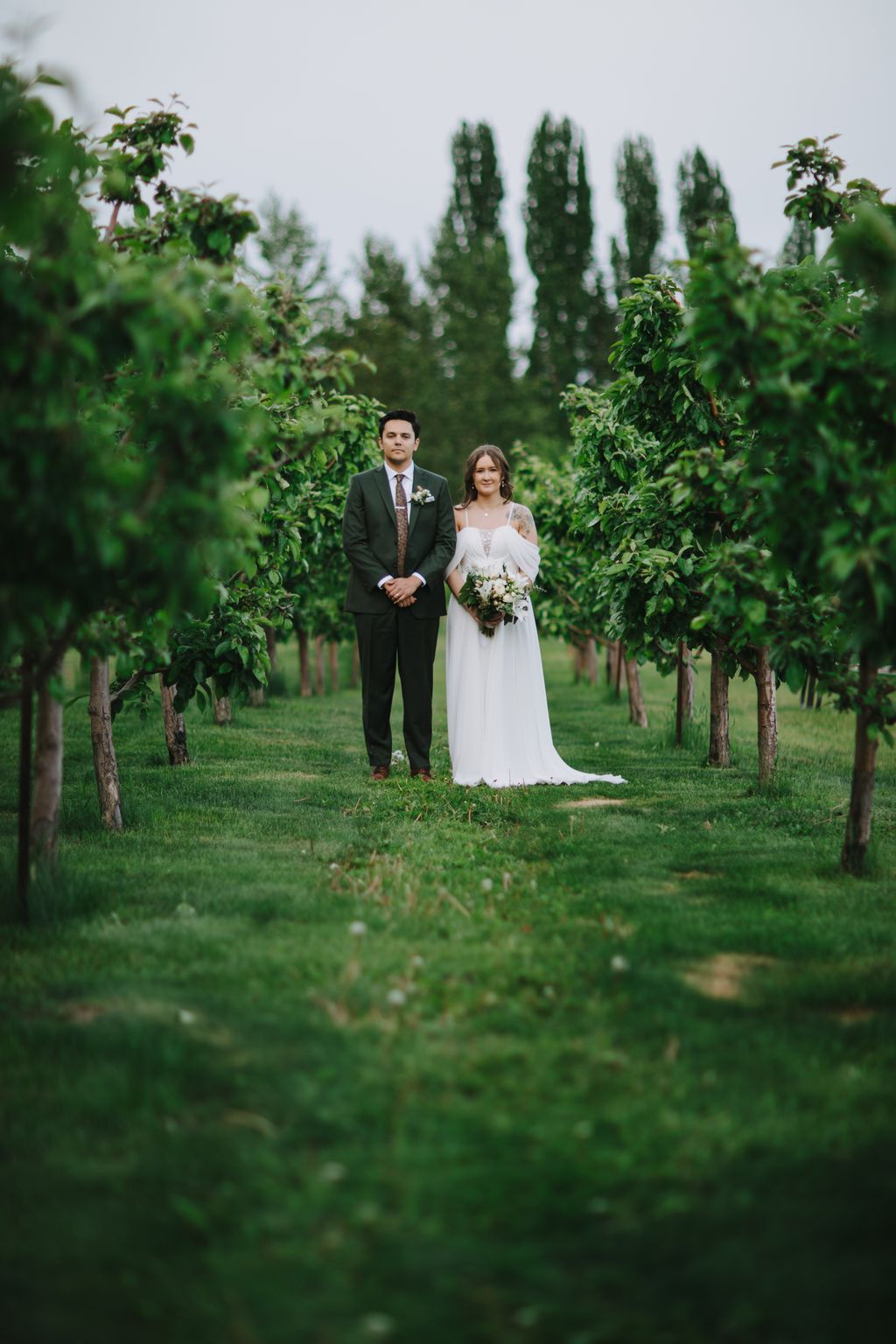 Bride and groom stand together in a lush garden corridor lined with tall green hedges under overcast skies