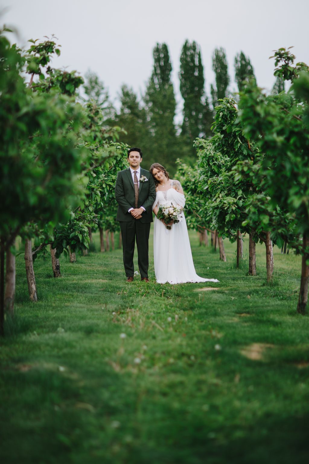 Bride in white off-shoulder gown and groom in charcoal suit stand together in a vineyard with tall cypress trees, holding hands and looking at camera