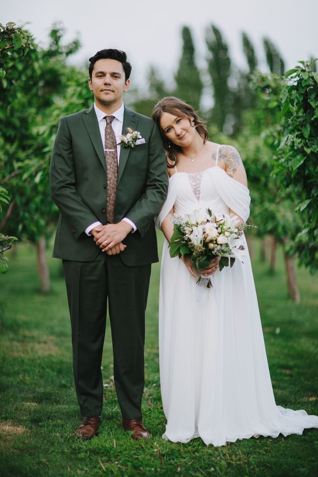 Bride in white dress holding wildflower bouquet stands beside groom in green suit against cypress trees and soft overcast sky