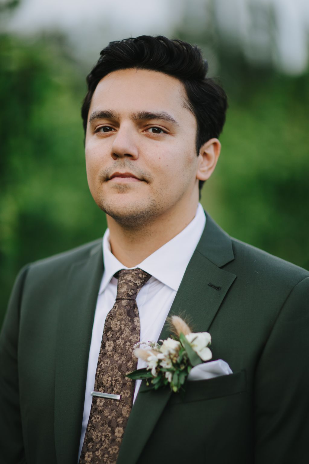 Groom in sage green suit and patterned tie with white boutonniere, soft outdoor portrait with blurred green background