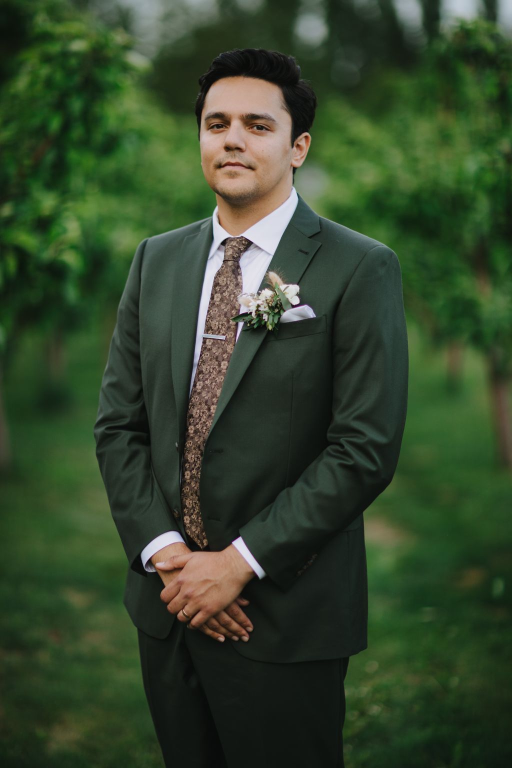 Groom in sage green suit with patterned tie and boutonniere stands against blurred green garden backdrop