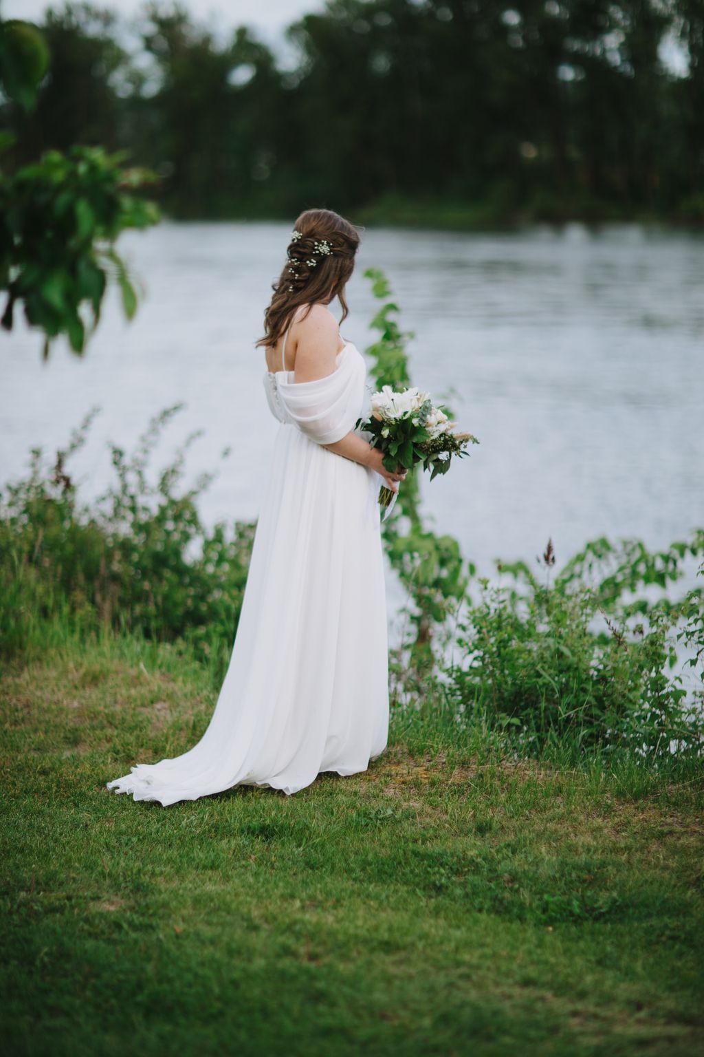 Bride in flowing white off-shoulder gown holding white bouquet, standing by riverside with green foliage and golden hair accessories