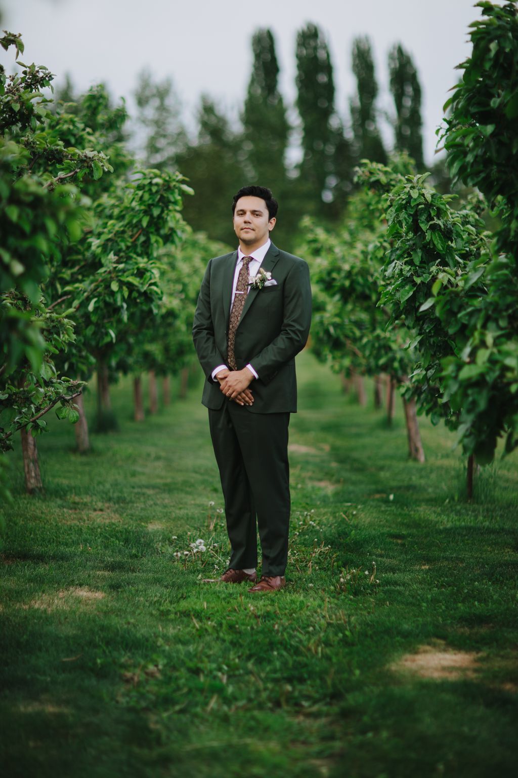 Groom in charcoal suit and floral tie stands in lush garden with cypress trees, wearing boutonniere