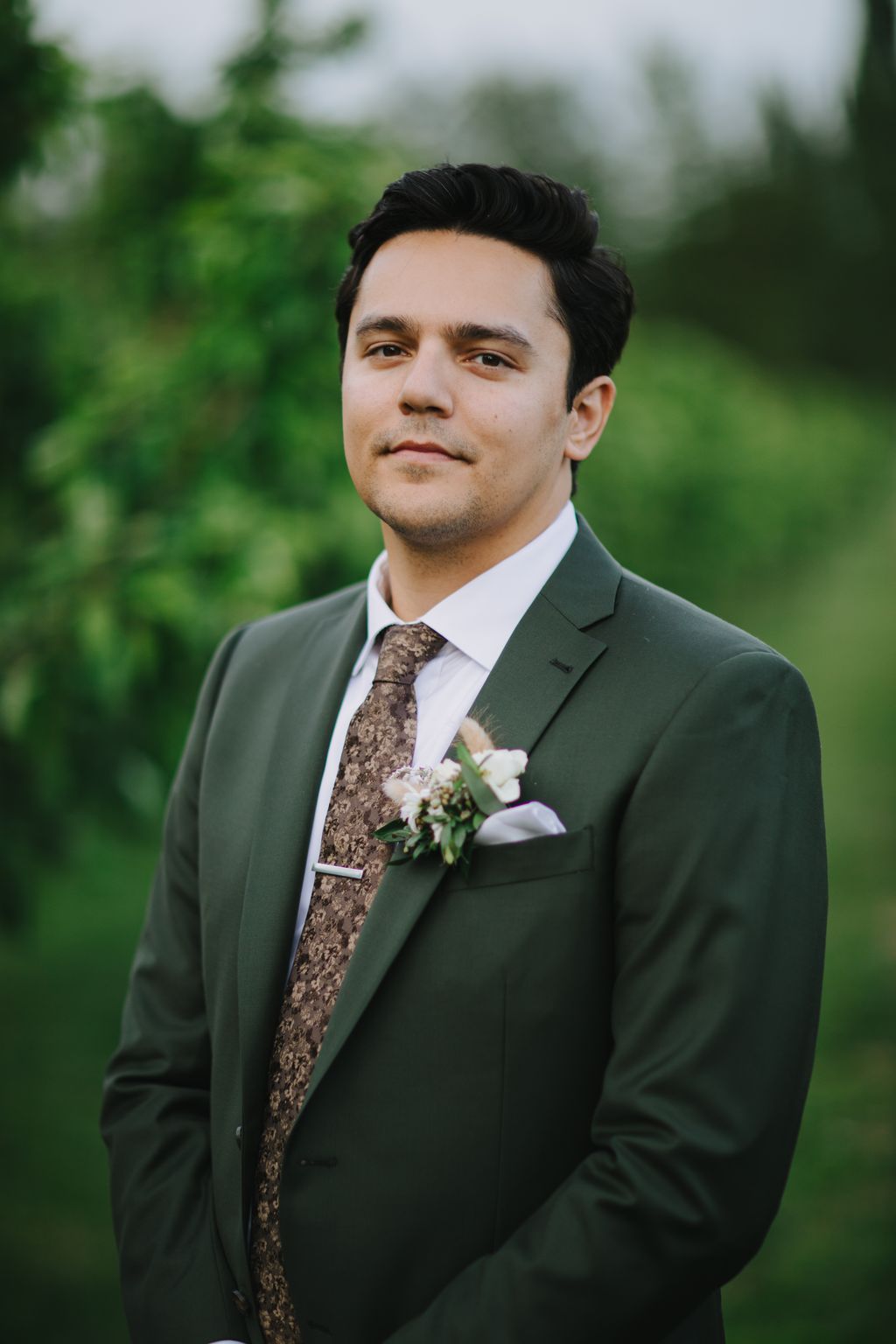 Groom in sage green suit with patterned tie and boutonniere poses outdoors with soft, blurred green foliage background
