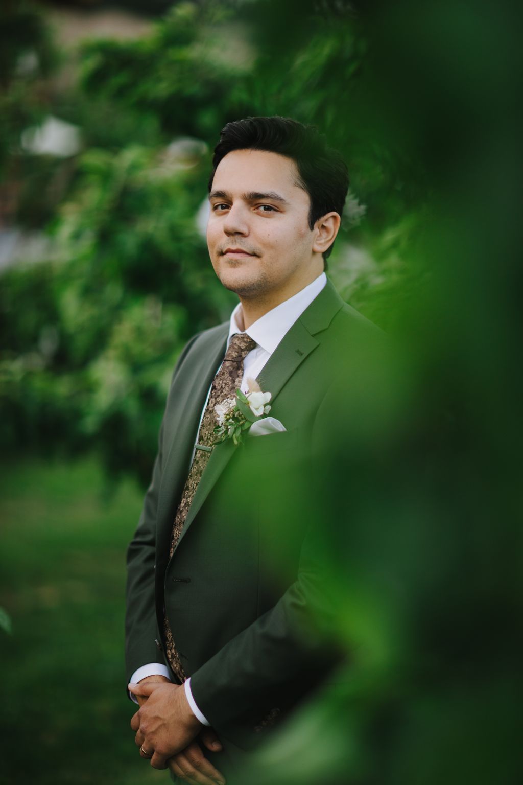 Groom in sage green suit and patterned tie with boutonniere poses against lush green foliage backdrop