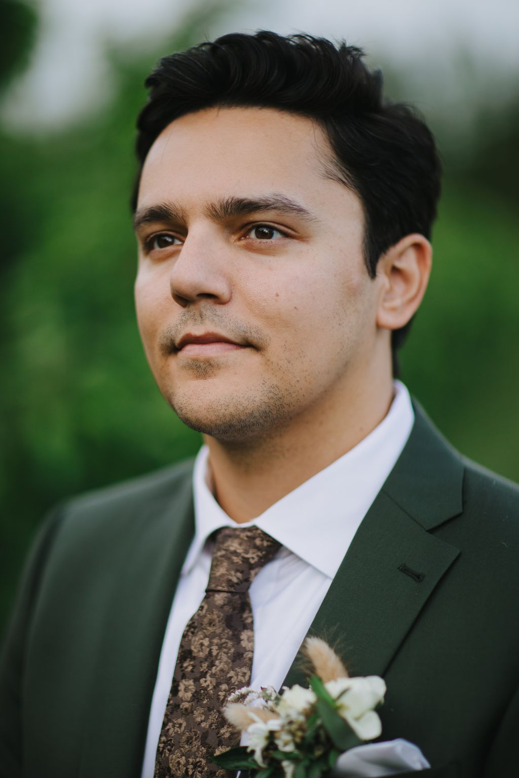 Groom in grey suit and brown patterned tie with white boutonniere, soft natural light, blurred green background