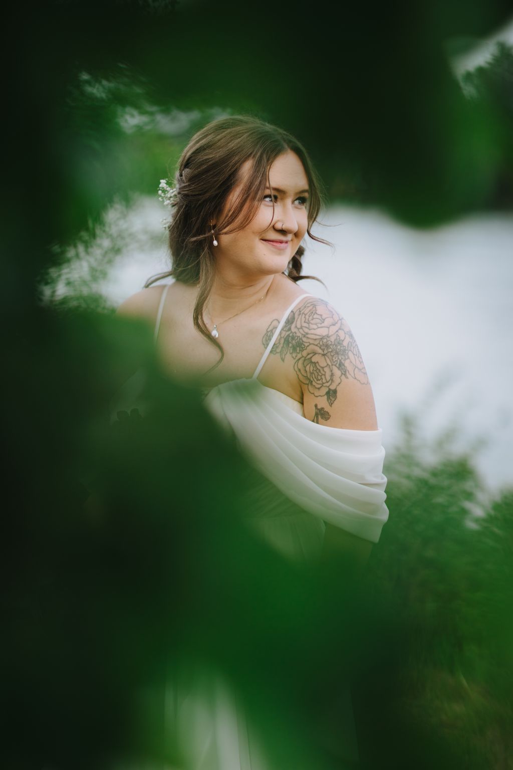 Bride with floral shoulder tattoo in white draped fabric, styled hair with baby''s breath, smiling softly against blurred green foliage backdrop