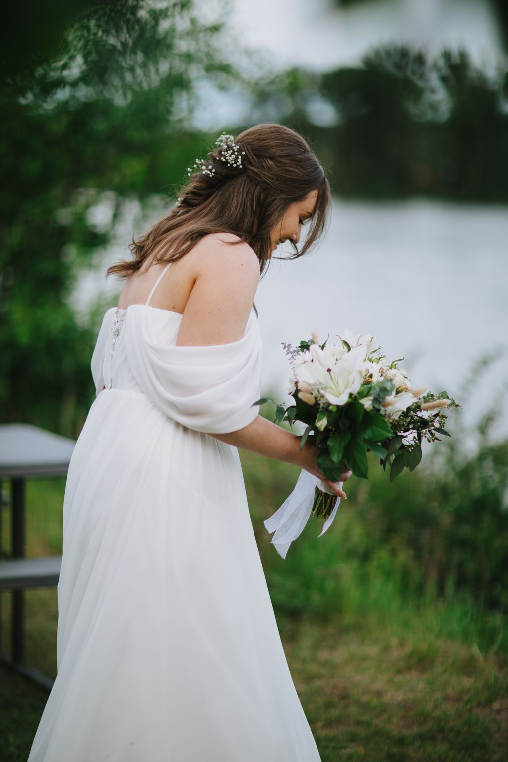 Bride in flowing white dress holding white flower bouquet, wearing baby''s breath hair clip, overlooking garden setting with soft natural light