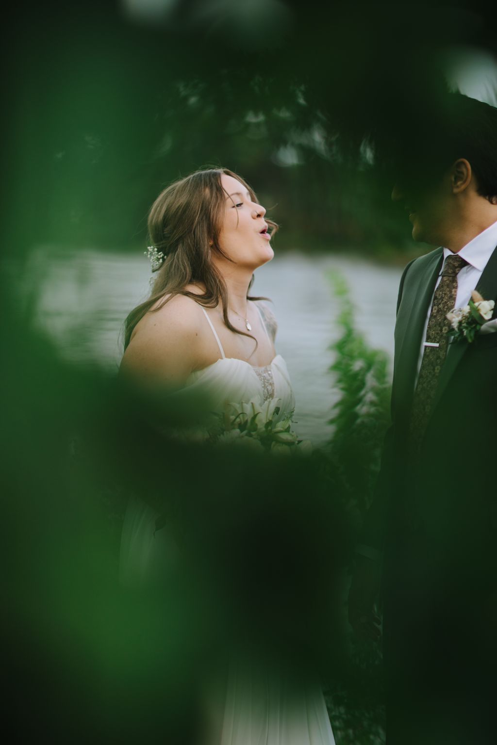 Bride in delicate white gown with pearl hair accessory gazes upward, surrounded by soft green foliage in natural light