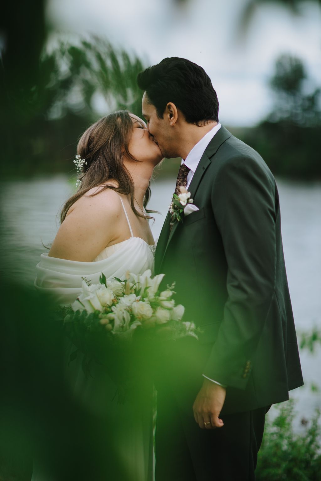 Bride and groom kiss outdoors with white flowers and greenery, soft natural light, tropical setting
