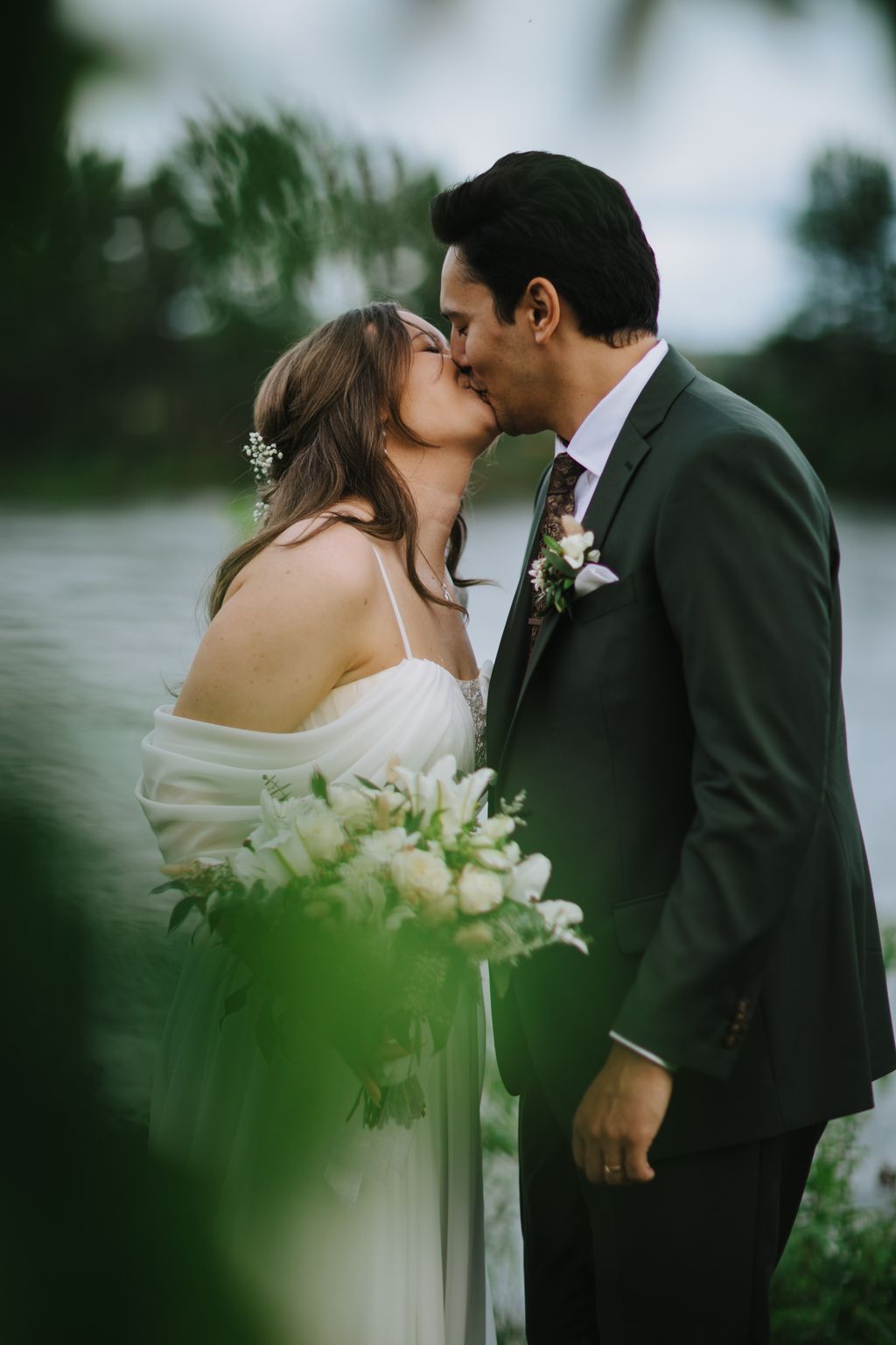 Bride and groom kiss outdoors with white flowers and lush green foliage in soft, natural light
