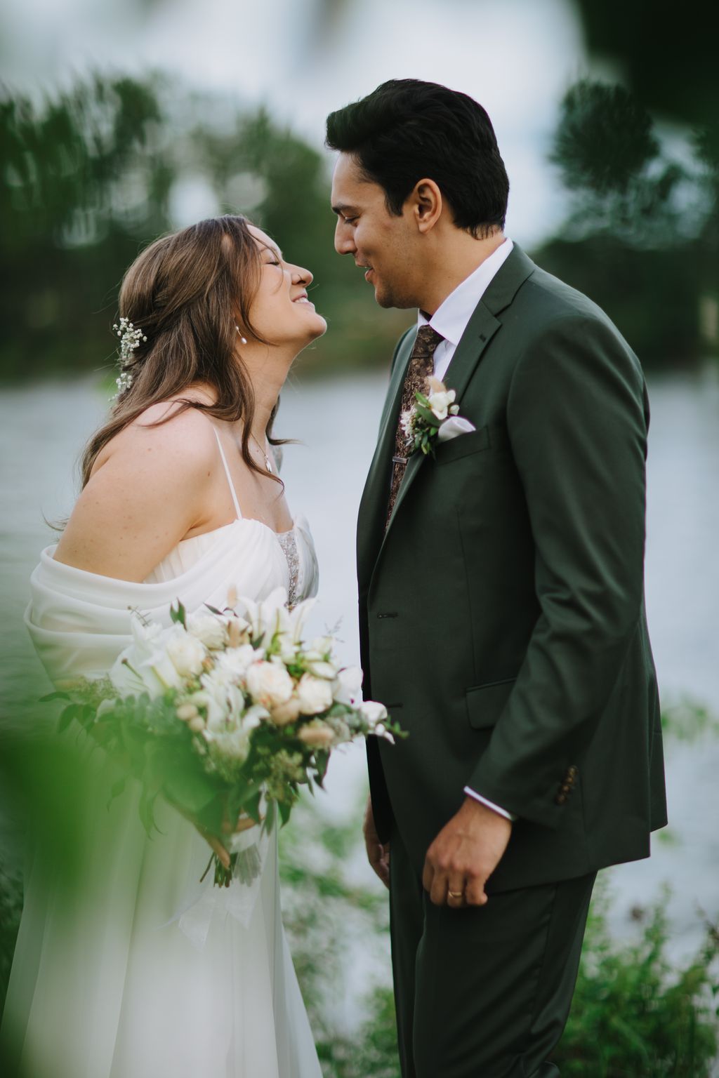 Bride and groom face each other smiling outdoors, she holding white flowers, he wearing sage green suit, with blurred water and trees behind