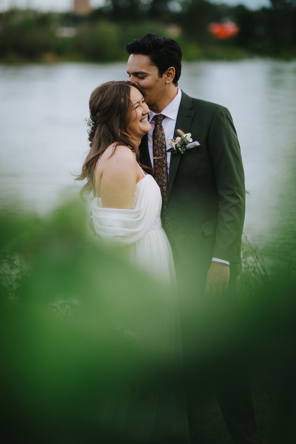 Groom in dark green suit kisses bride''s forehead while she smiles, outdoor garden setting with soft natural light and blurred greenery