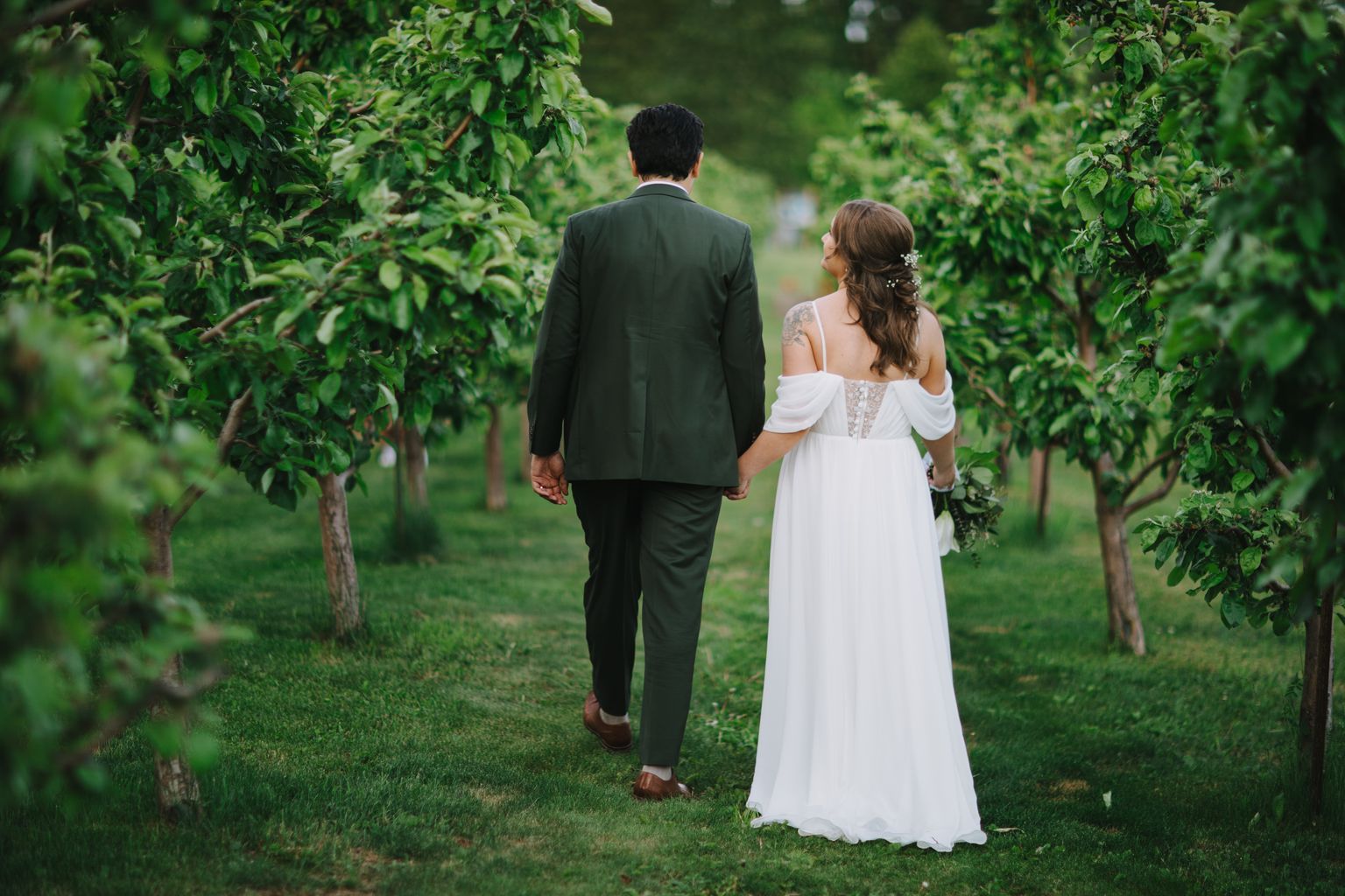 Bride and groom walk hand in hand through a green garden, the groom in a dark suit and the bride in a white off-shoulder gown with lace details