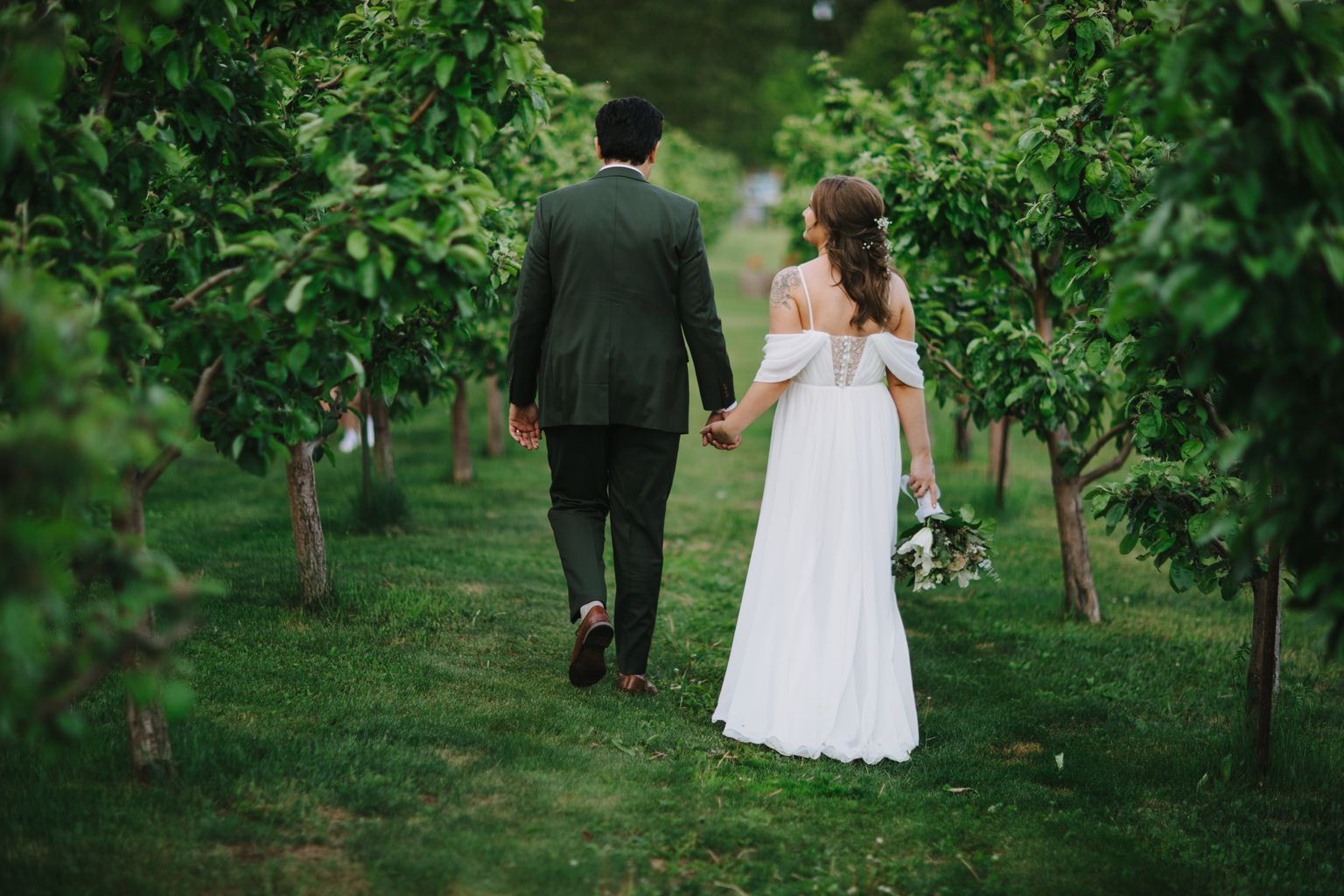 Bride and groom holding hands walking through lush green garden, bride in white off-shoulder gown, groom in dark suit