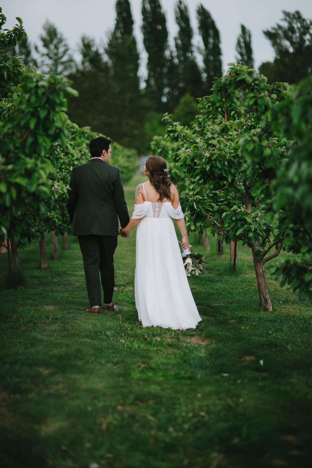 Bride in white off-shoulder gown and groom in dark suit hold hands walking through lush garden with tall cypress trees