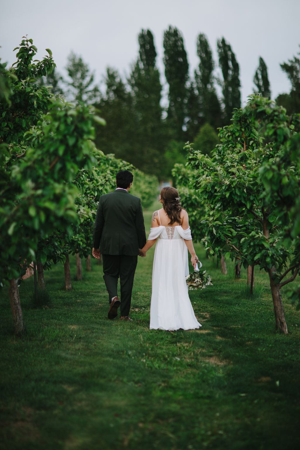 Bride and groom holding hands walking through a lush garden with tall cypress trees and green hedges, overcast daylight
