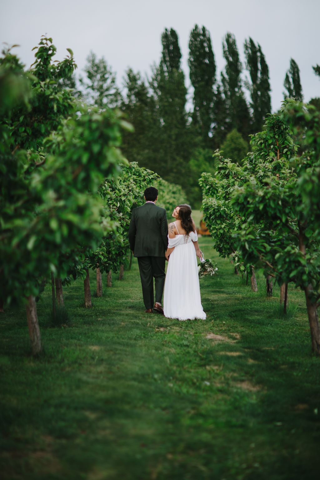 Bride in white off-shoulder gown and groom in black suit walking hand-in-hand through lush green vineyard rows with tall cypress trees in soft, overcast light