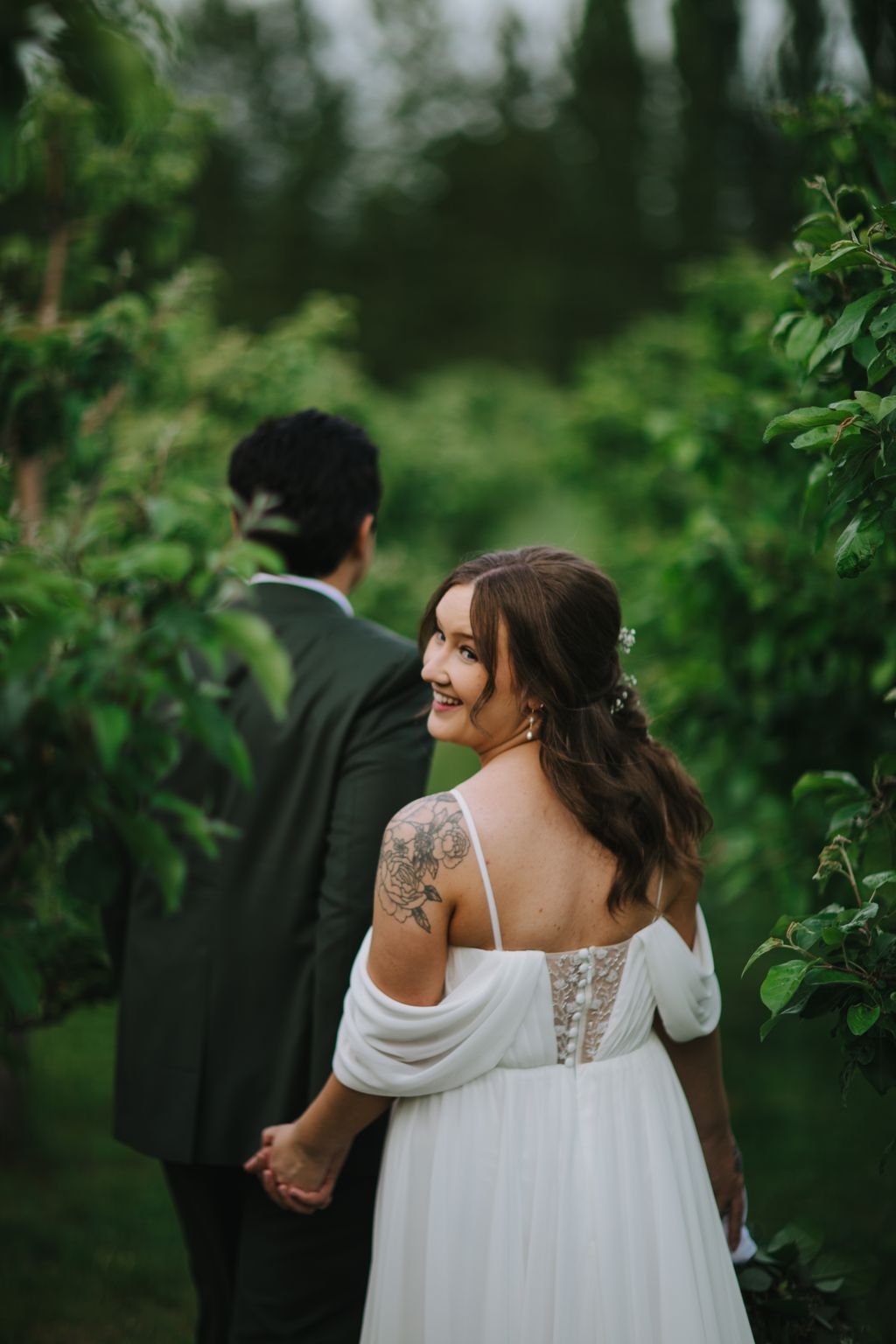 Bride in flowing white dress with floral shoulder tattoo smiles over her shoulder while holding groom''s hand in lush garden setting