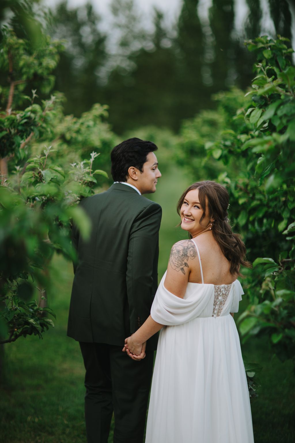 Bride and groom holding hands in a lush garden, smiling with evergreen trees softly blurred in background
