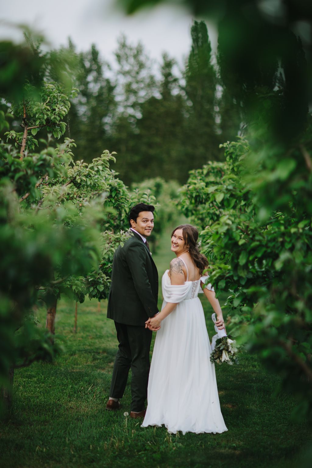 Bride and groom holding hands in a lush green garden, surrounded by flowering shrubs and tall evergreens under overcast skies