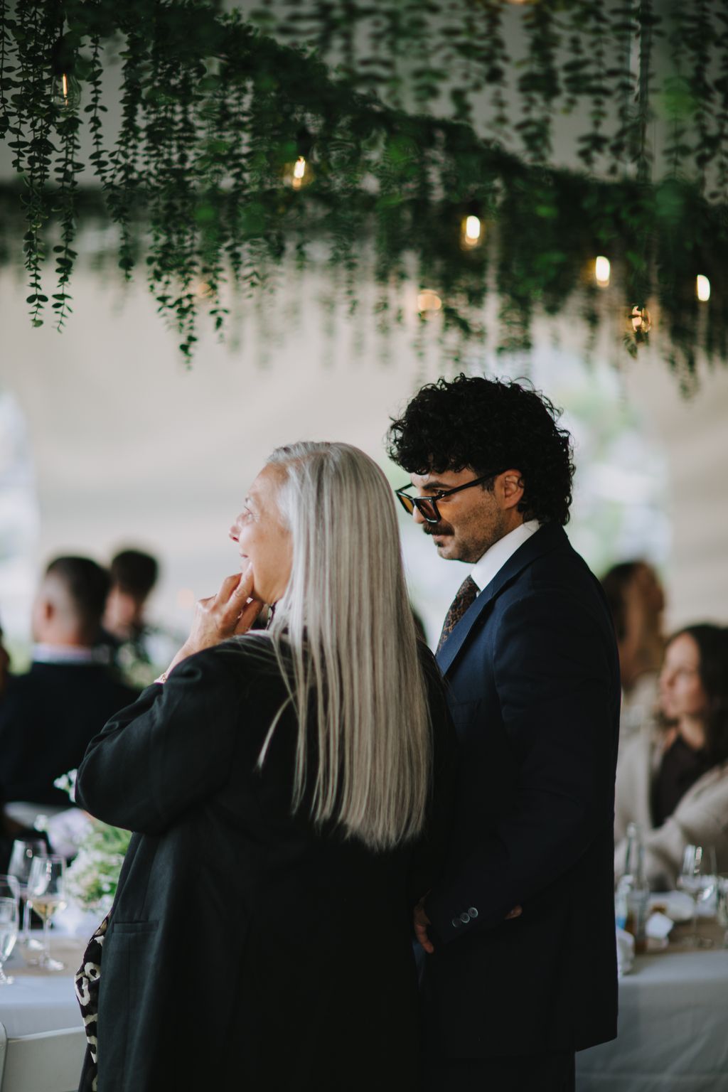 Couple in formal attire stands beneath cascading greenery and warm pendant lights in an elegant indoor venue