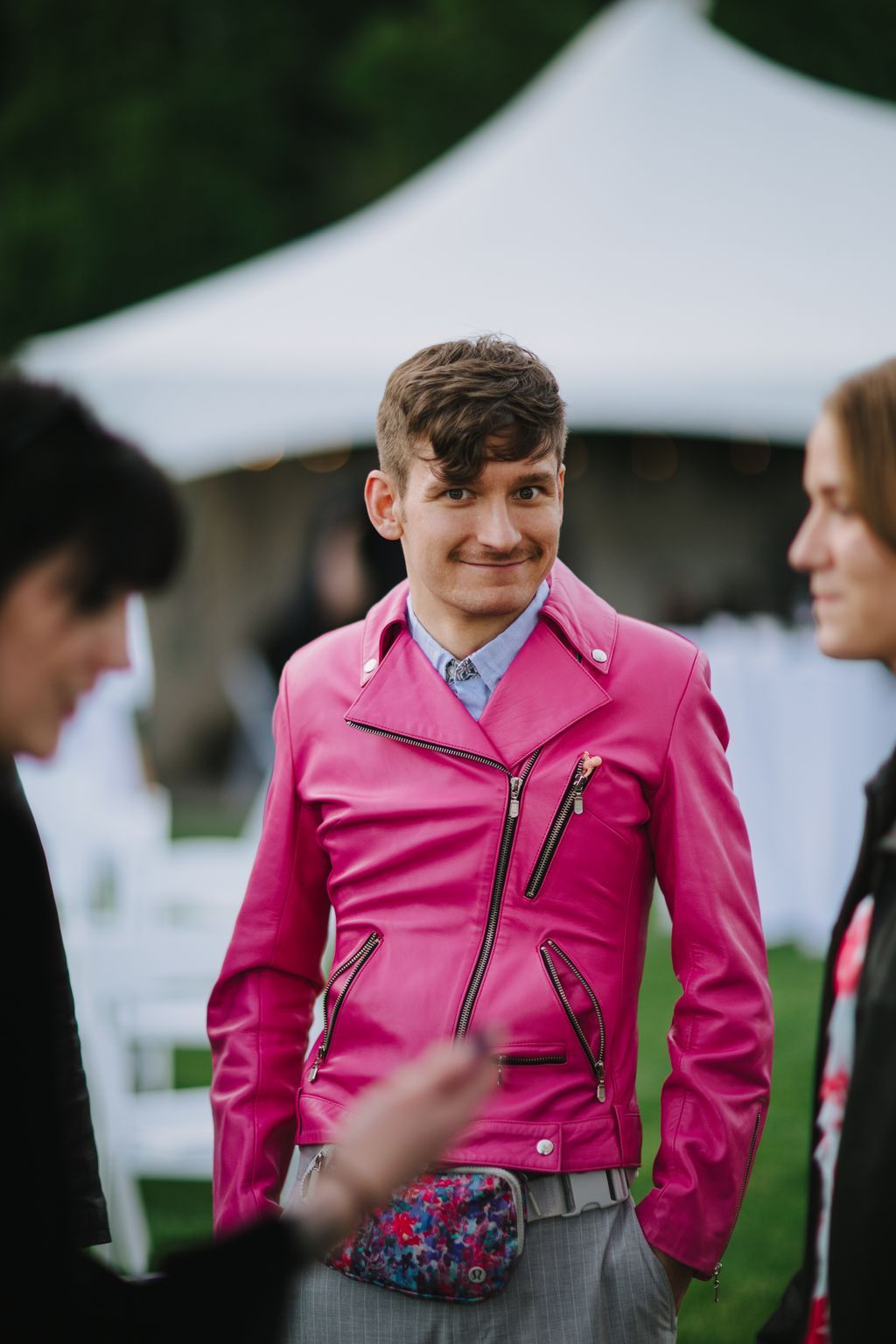 Groom in bright pink jacket with blue shirt smiles at outdoor venue with white tent backdrop