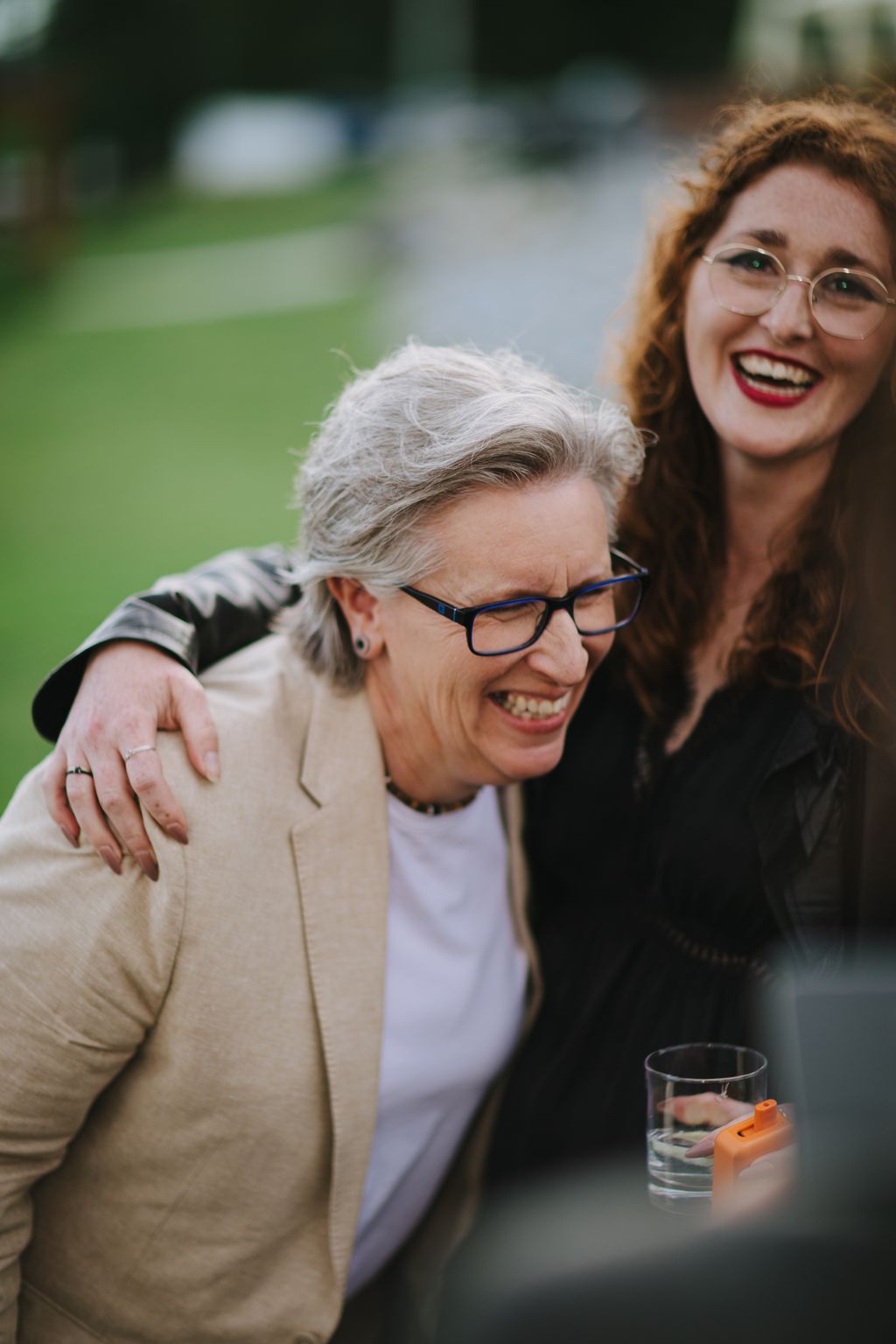 Smiling woman with gray hair and glasses wearing a beige blazer at an outdoor garden celebration
