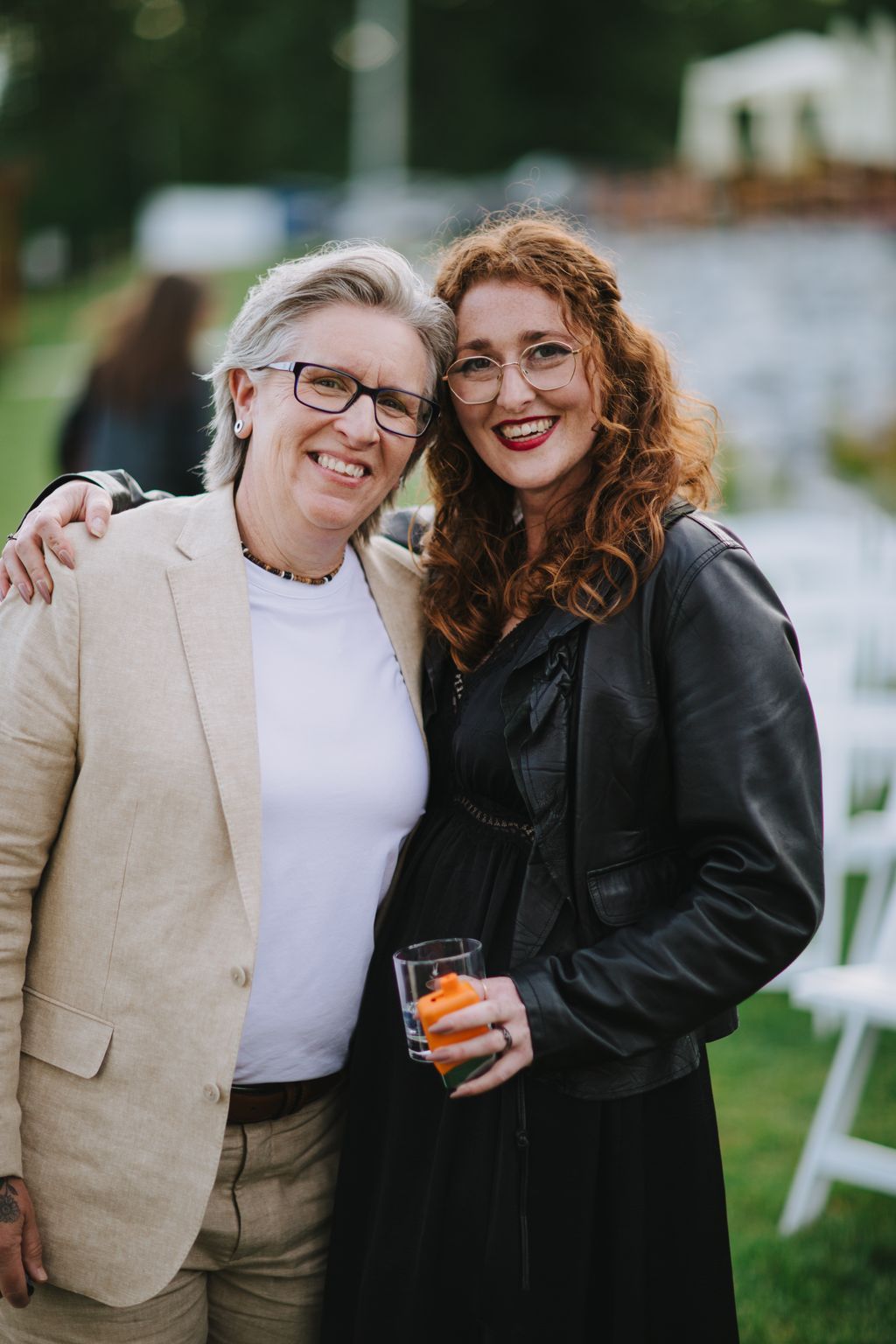 Two women smiling at an outdoor event, both wearing glasses and holding drinks, with blurred green background