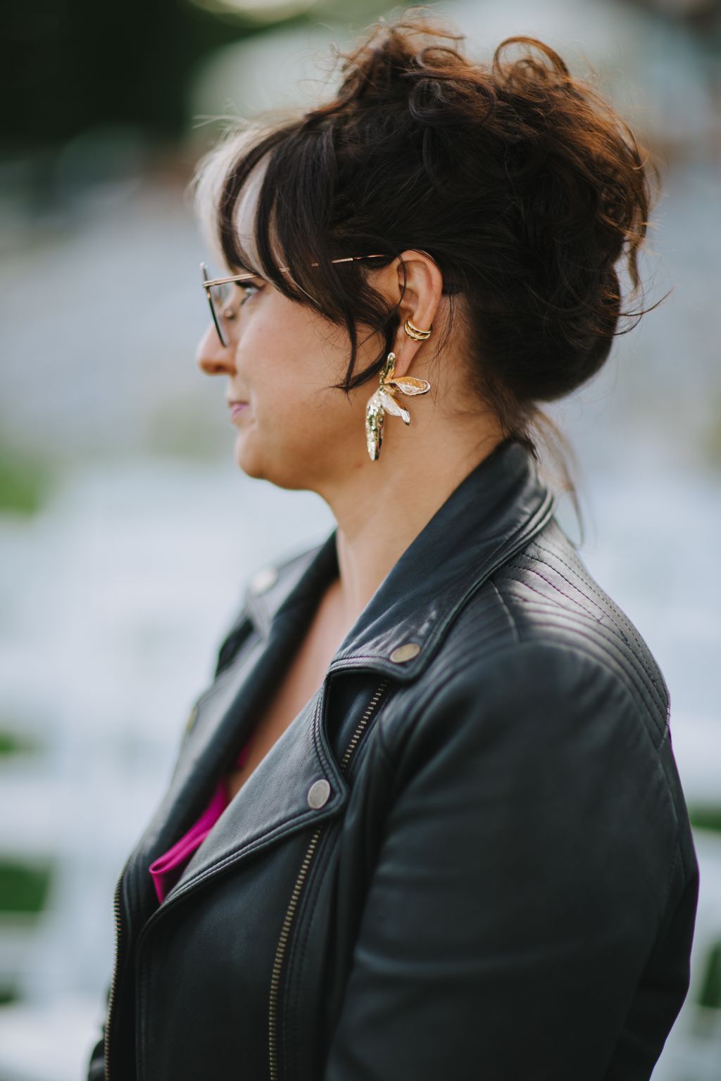 Woman in profile wearing glasses and statement leaf earrings, black leather jacket over magenta top, soft natural light