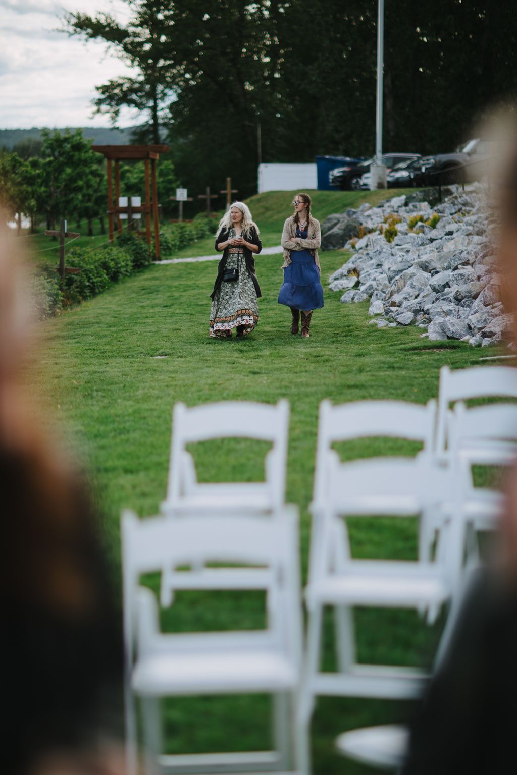 Two women walk across a grassy lawn with white chairs blurred in the foreground during an outdoor ceremony