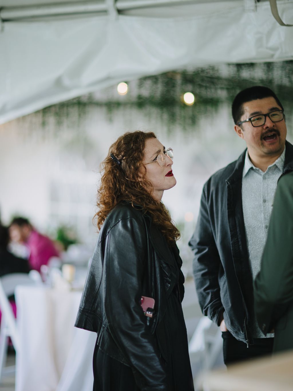 Woman in glasses and black leather jacket stands under white tent with greenery and warm lights, looking upward