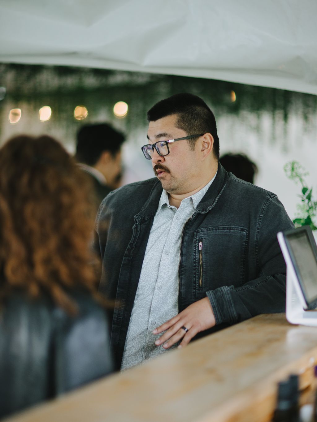 Man in glasses and denim jacket at indoor event with blurred background and warm ambient lighting