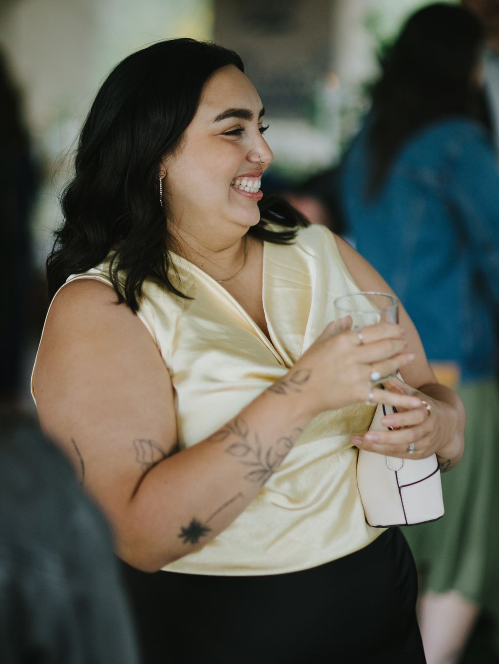 Woman in pale yellow shirt with visible tattoos smiles warmly in soft natural light, outdoor setting