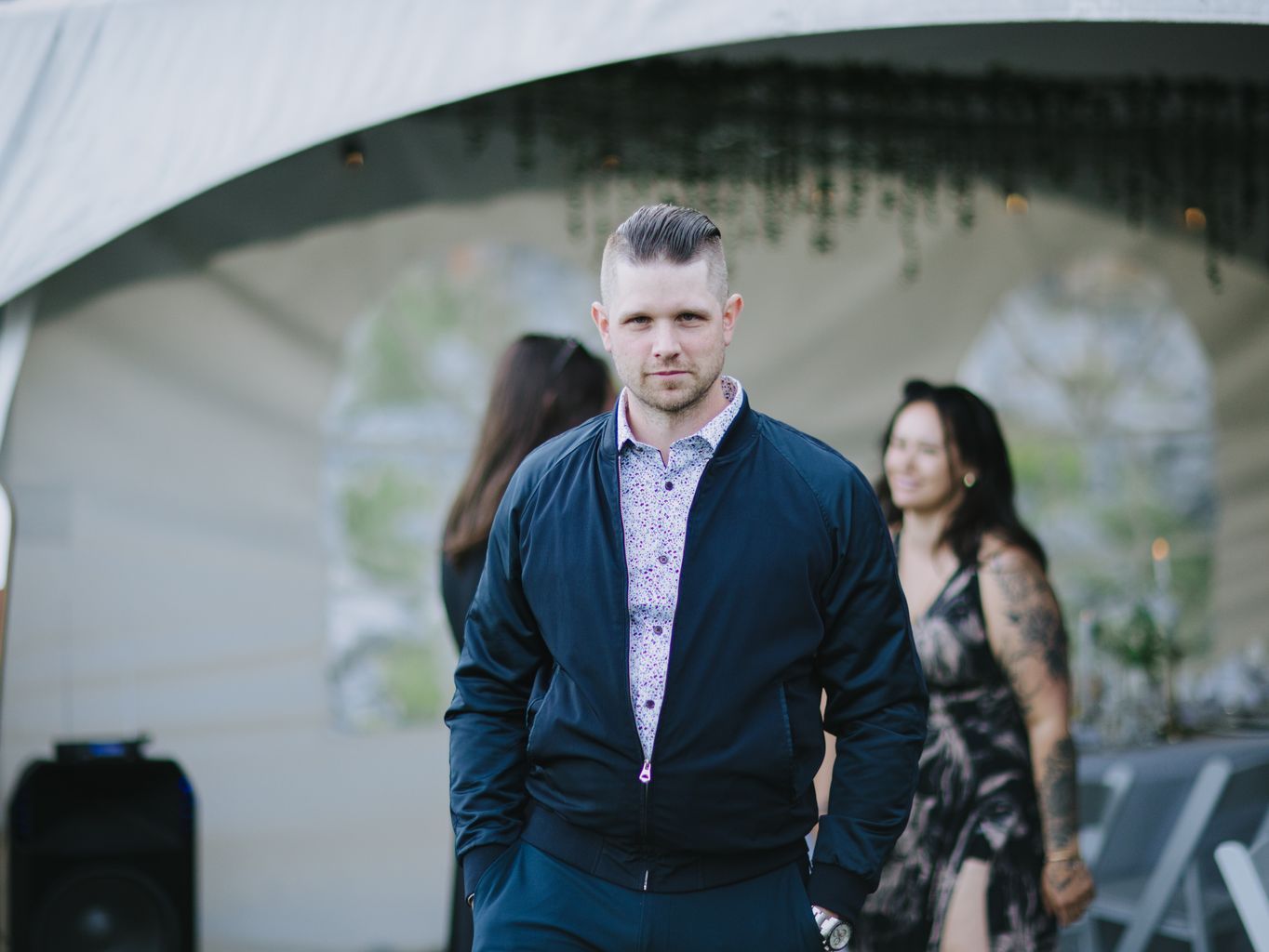 Man in navy jacket and patterned shirt stands outdoors with blurred figure in background against textured wall