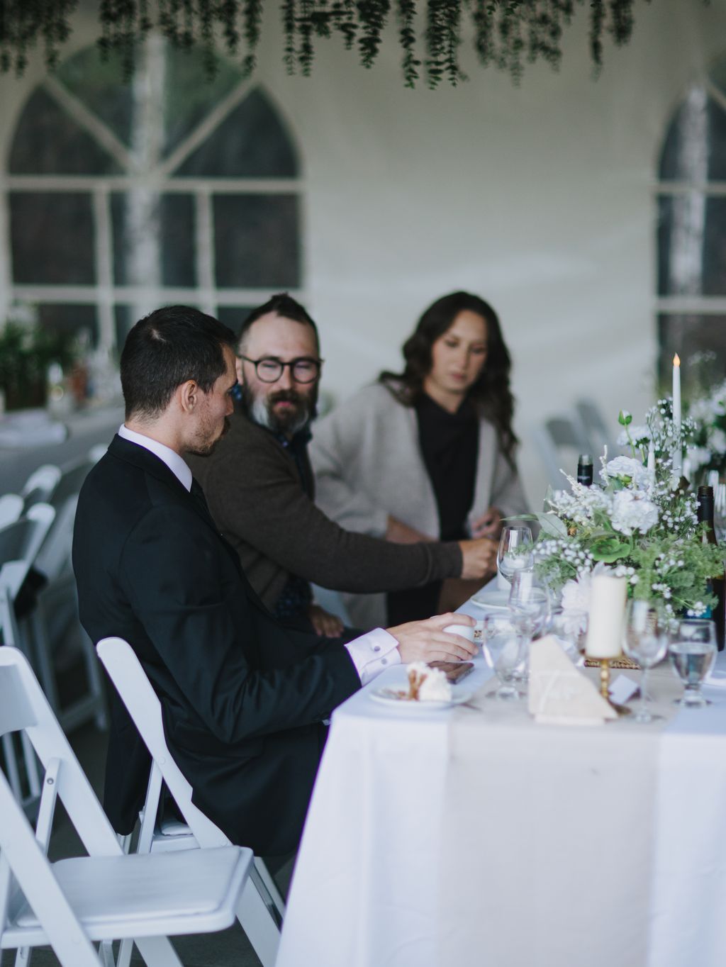 Three people seated at a white-draped table during an indoor reception, with hanging greenery and soft natural light