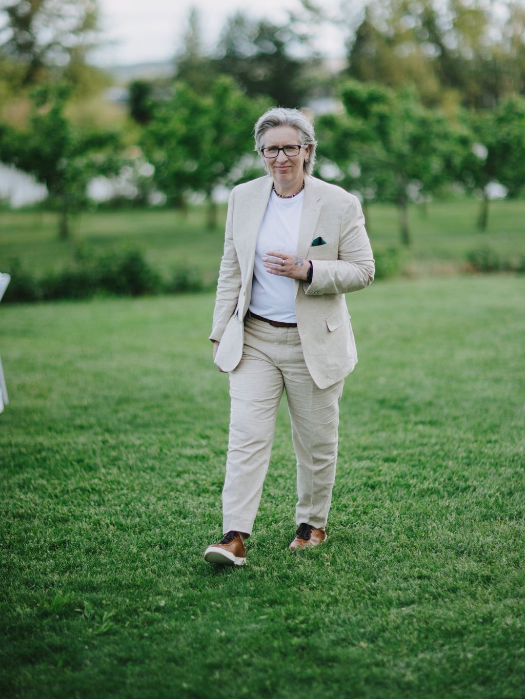 Woman in cream linen suit and glasses standing on grass with blooming garden backdrop, soft natural light