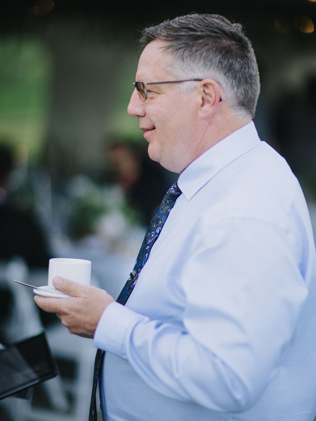 Groom in light blue shirt and patterned navy tie smiles in profile during outdoor ceremony