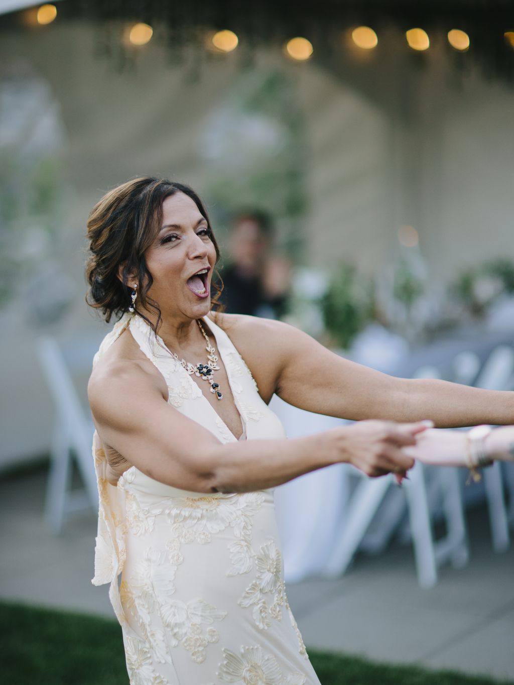 Woman in ivory beaded halter wedding dress laughs joyfully at outdoor evening reception with warm string lights glowing softly behind her