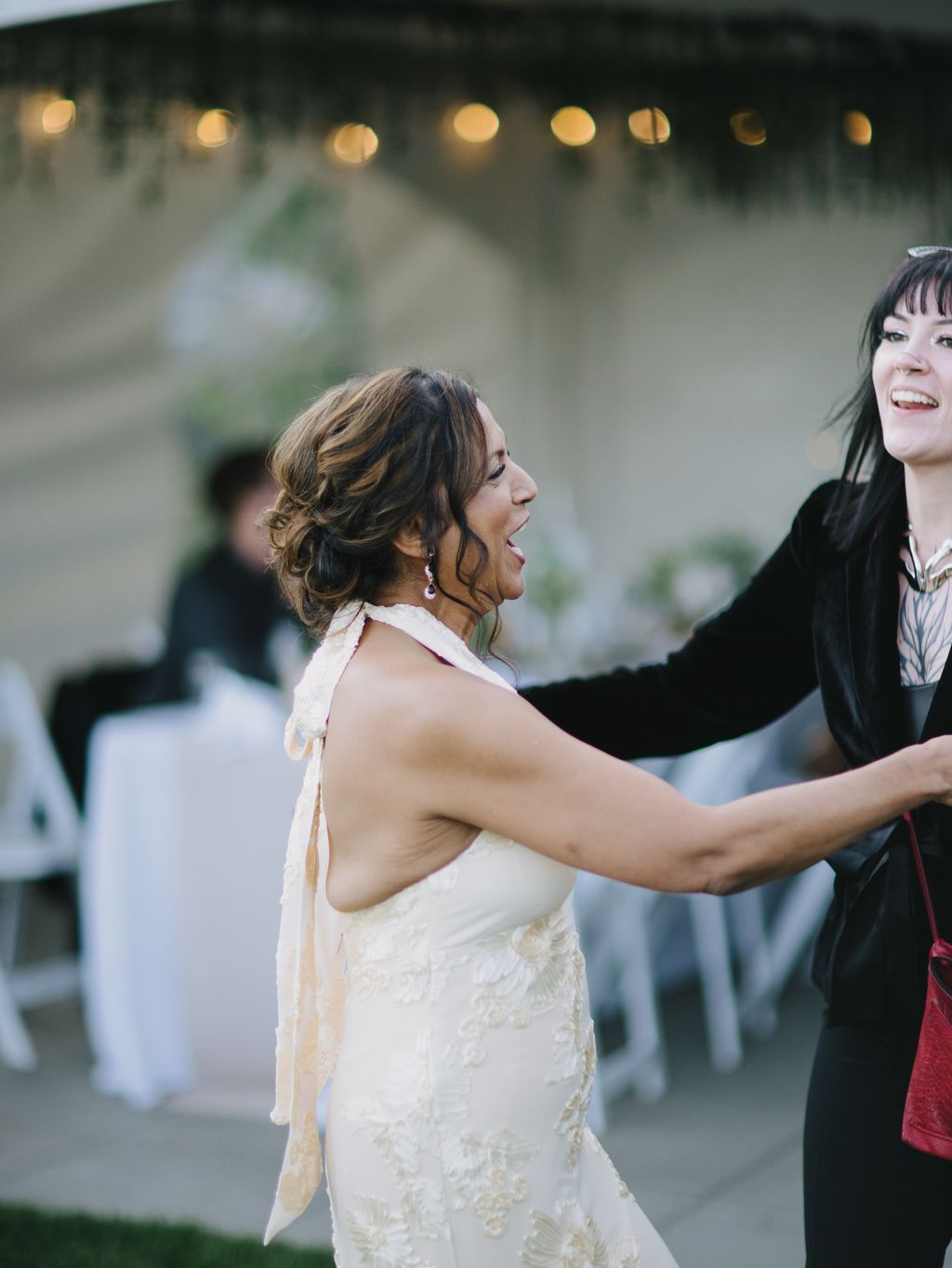 Bride in embellished white halter dress laughs with arms outstretched at evening reception under warm string lights
