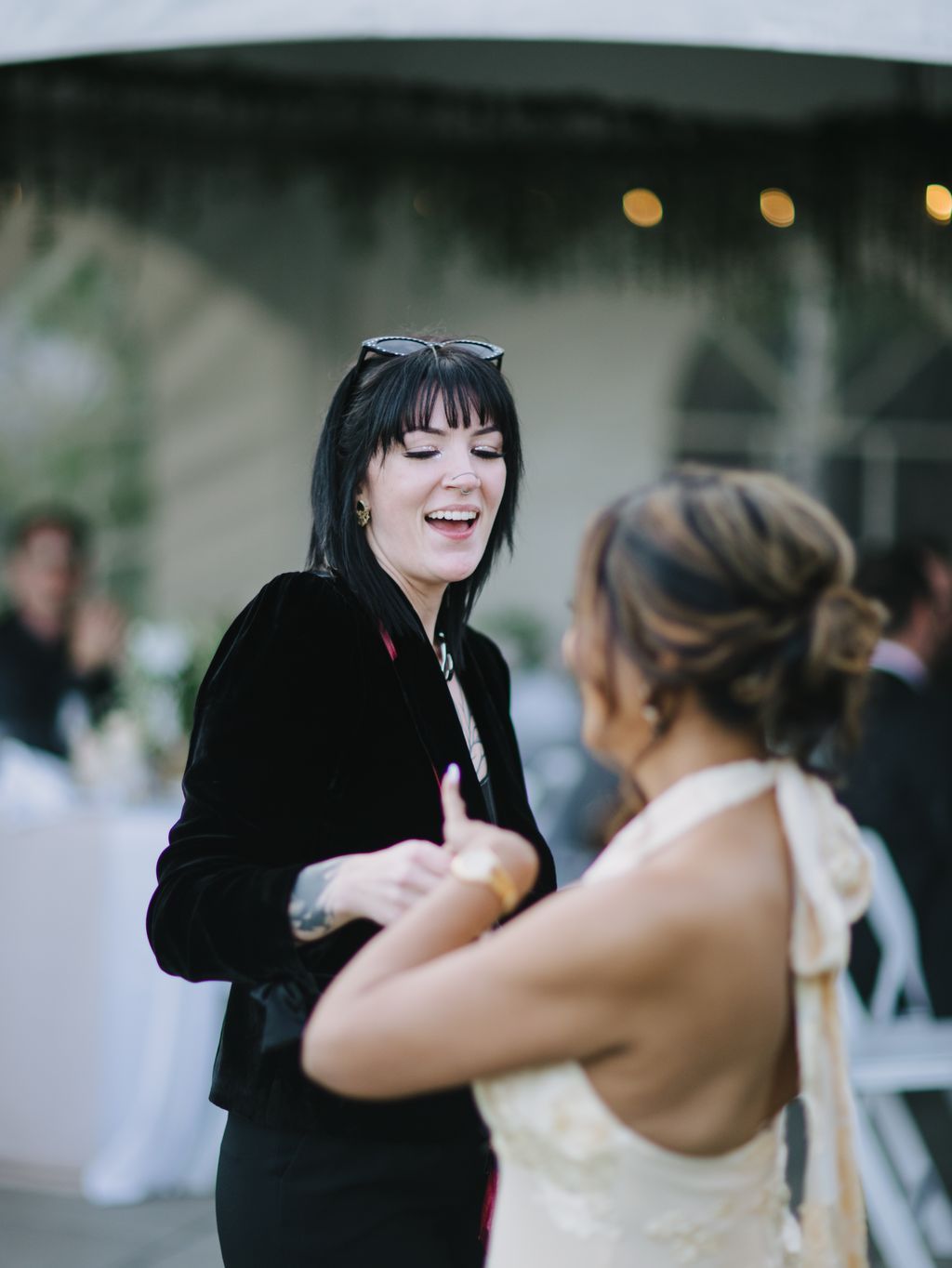 Woman in black blazer with sunglasses on head laughs while talking with another person outdoors at dusk