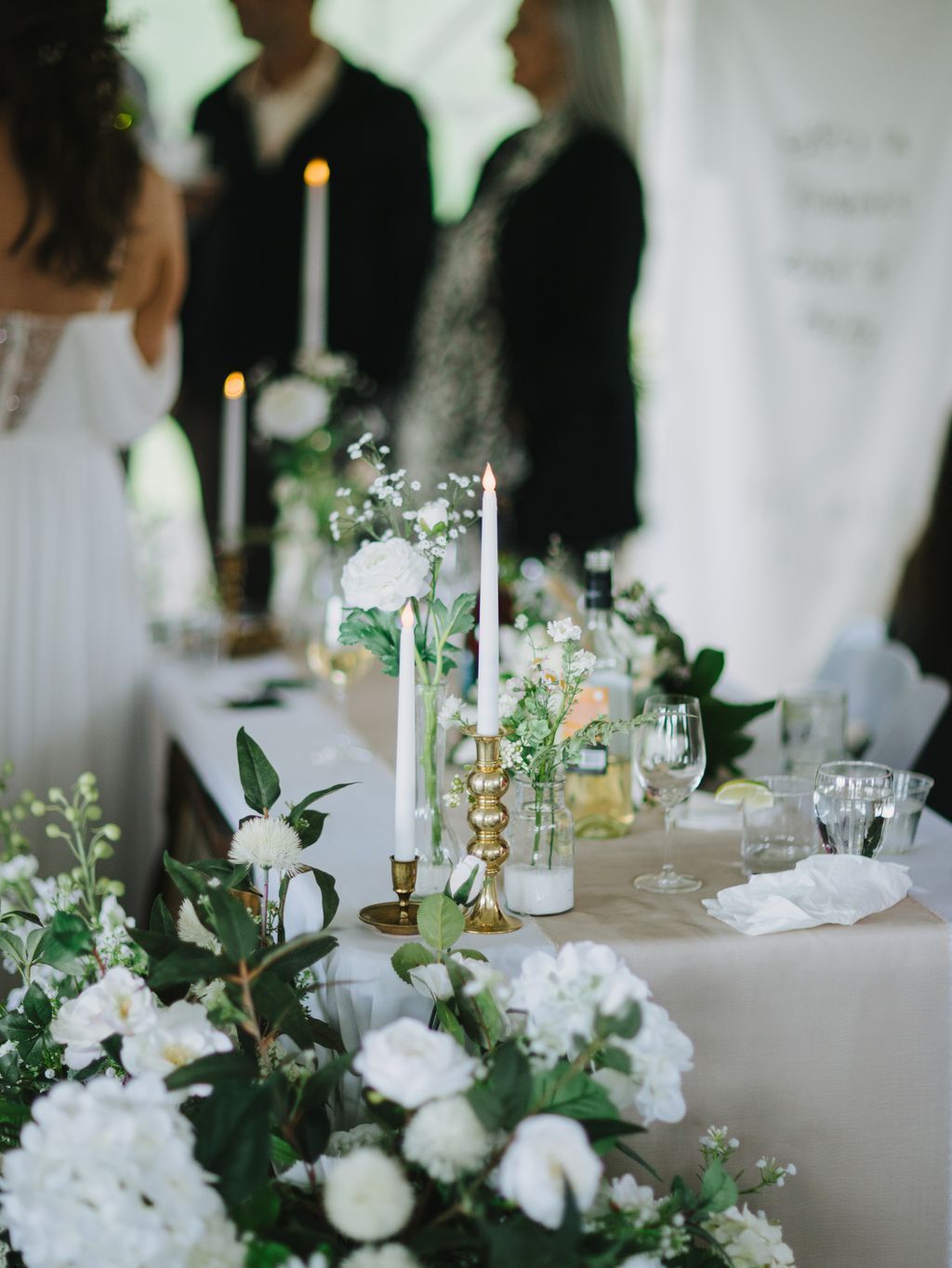 Elegant white table setting with white roses, baby''s breath, brass candlesticks, and lit candles, guests in dark clothing blurred in background