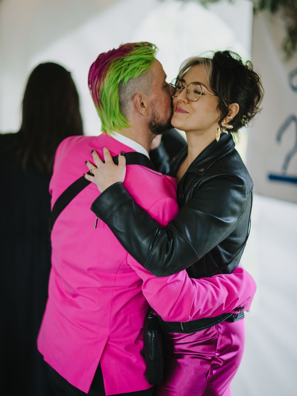 Two people embrace closely against a white background, one wearing a vibrant pink suit with lime green and magenta hair, the other in black leather and glasses