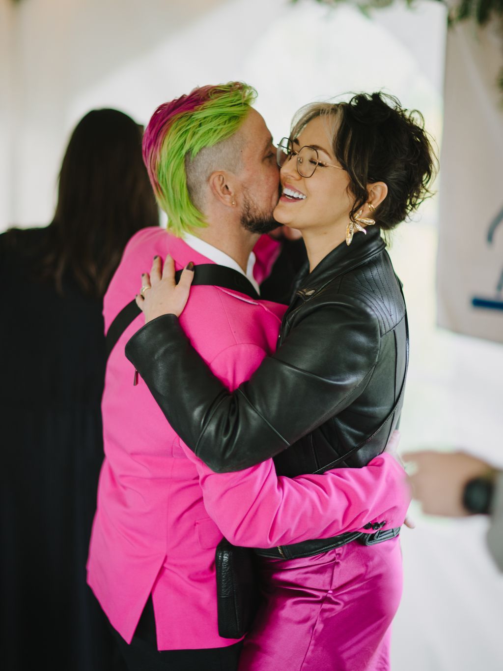 Two people in vibrant pink and black leather jackets share an affectionate kiss against a soft white backdrop, radiating joy and celebration