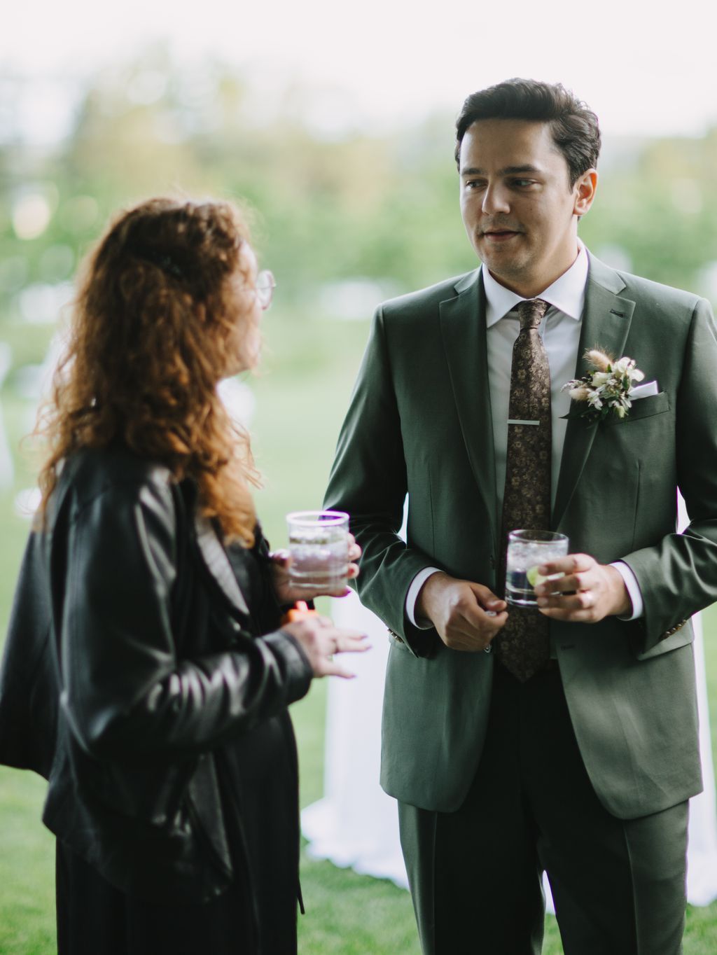 Groom in sage green suit converses with guest holding drink at outdoor reception with soft-focused greenery background