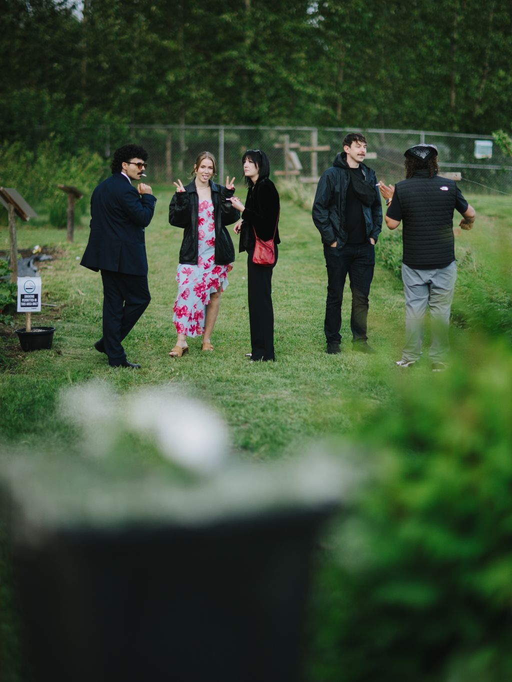 Four guests in dark formal attire stand on a grass field near a white cross, trees in soft focus behind them