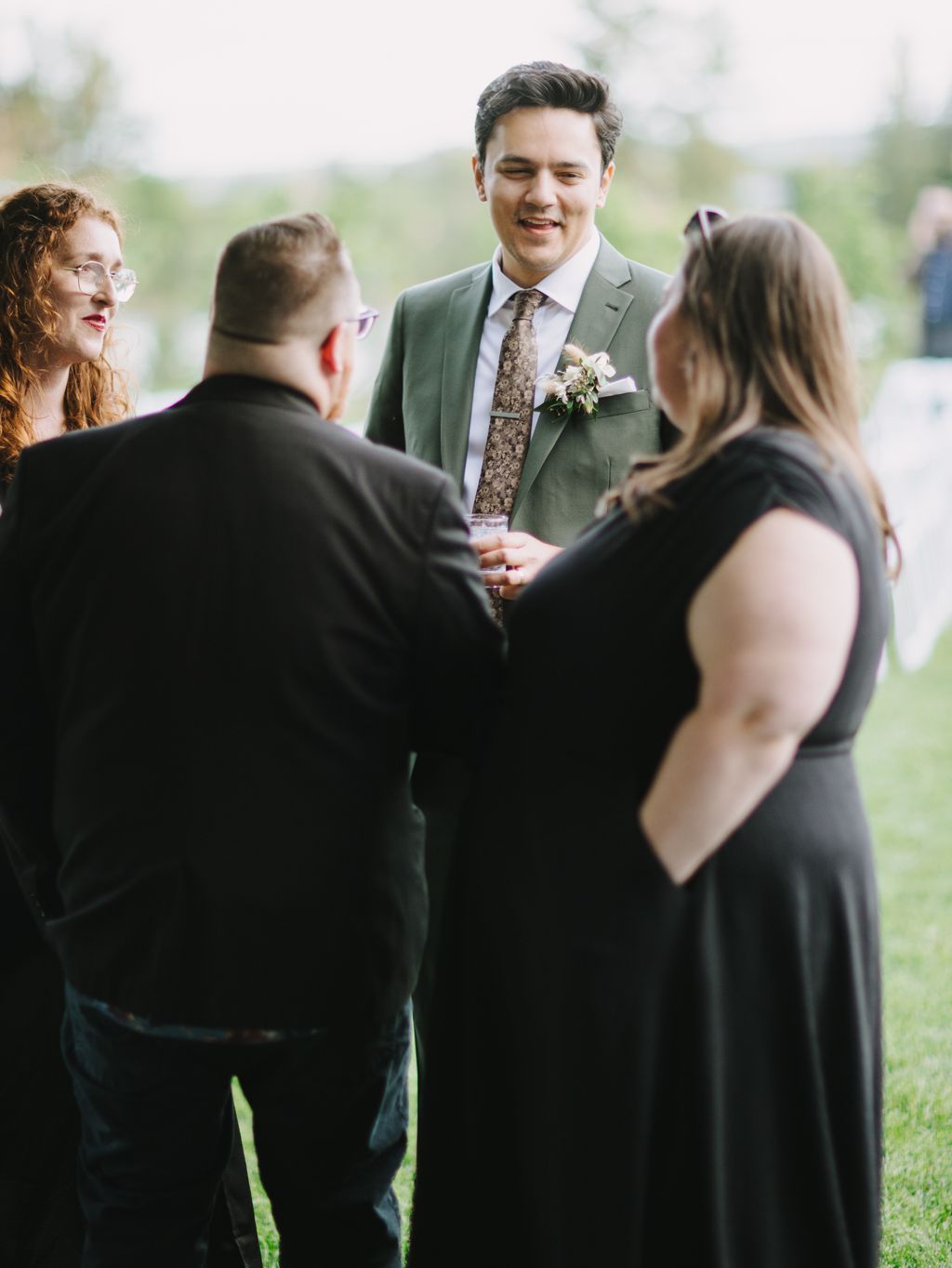 Groom in sage green suit smiles while conversing with guests outdoors on wedding day