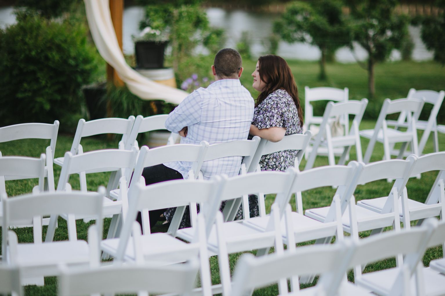 Couple seated in white chairs facing a garden, woman in floral dress and man in light blue shirt enjoying a quiet moment outdoors